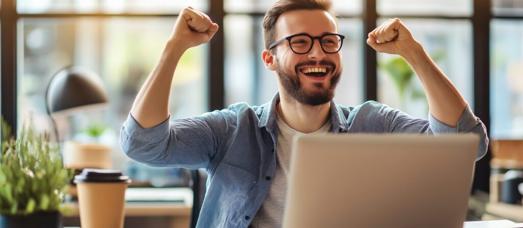 Happy young profitable freelance business man sitting in office at table, looking at laptop screen and enjoying achievement and success, making victory gesture with hand yes