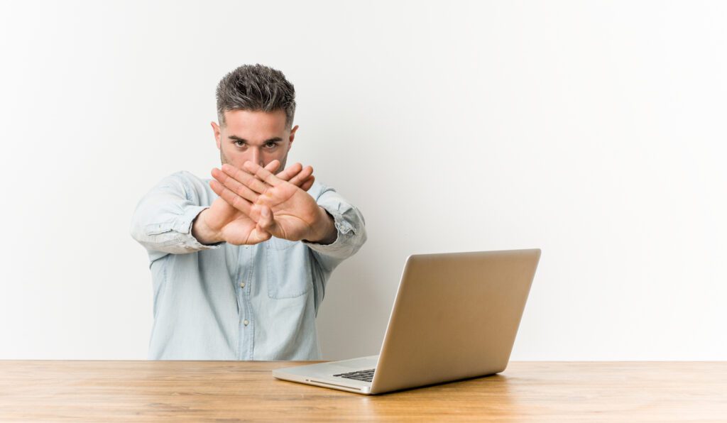 Young handsome man working with his laptop doing a denial gesture using the freelance productivity hacks "No"