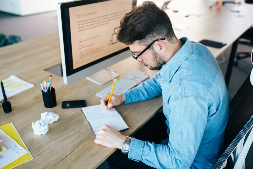 Young man in glassess is working at his workplace in office. He wears blue shirt.  He is writing in notebook. View from above.